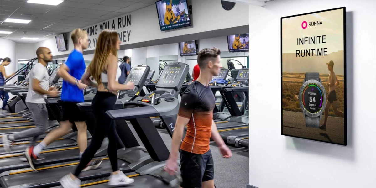 Digital screen in a gym displaying an ad for a smartwatch while people exercise on treadmills.