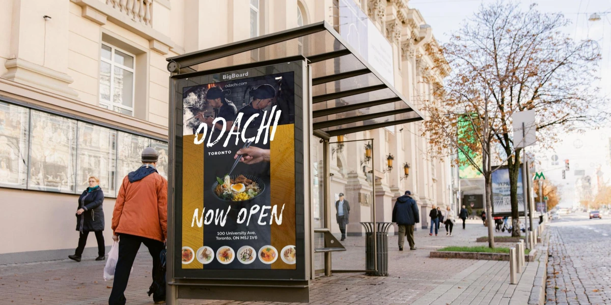 Digital bus shelter screen showing a DOOH ad for a ramen restaurant with the message 'Now Open'.