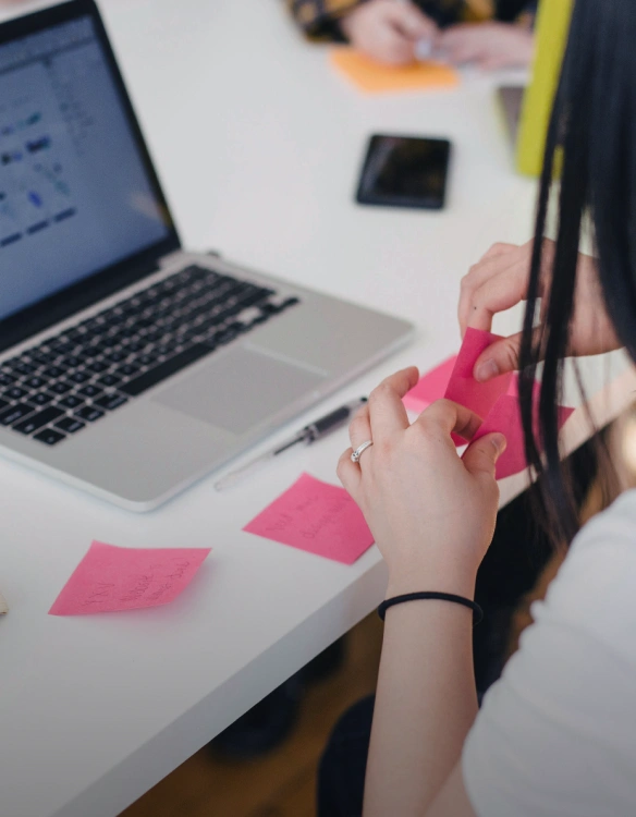 Individuals at a desk in front of laptops organizing and pasting sticky notes down with thoughts written out on them.
