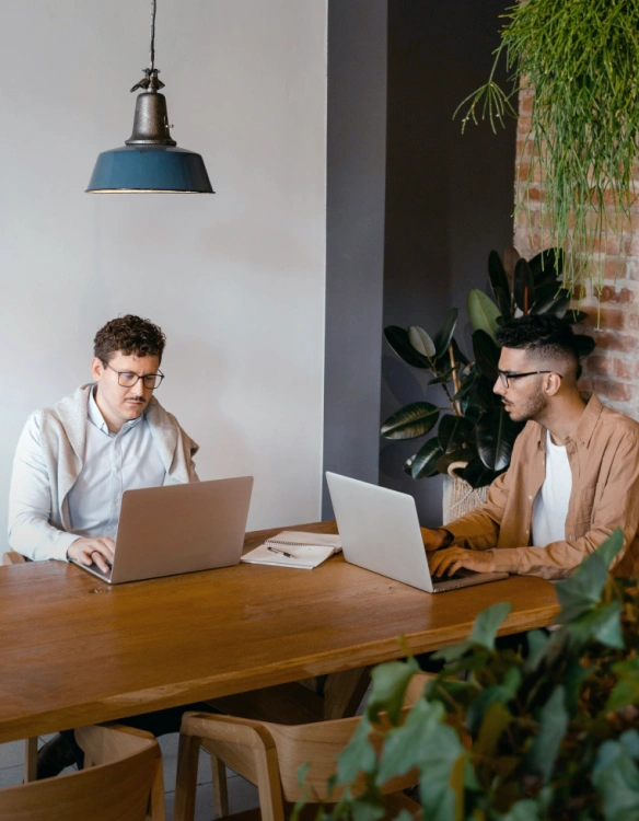 Two individuals seated at a large wooden desk with laptops open, both appear to be working on the devices while engaged in discussion.