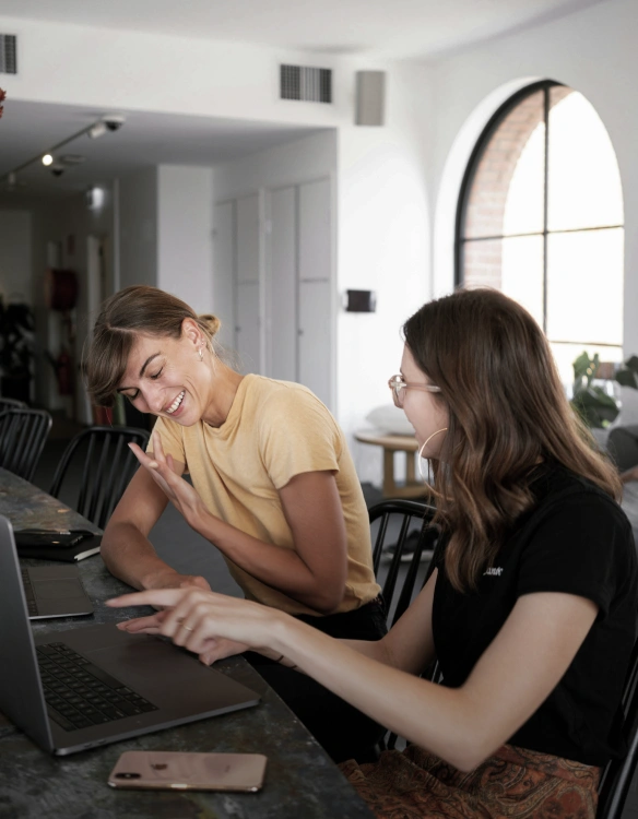 Two individuals seated at a desk with laptops in front of them, having a conversation about material displayed on the laptops suggestive of collaboration.