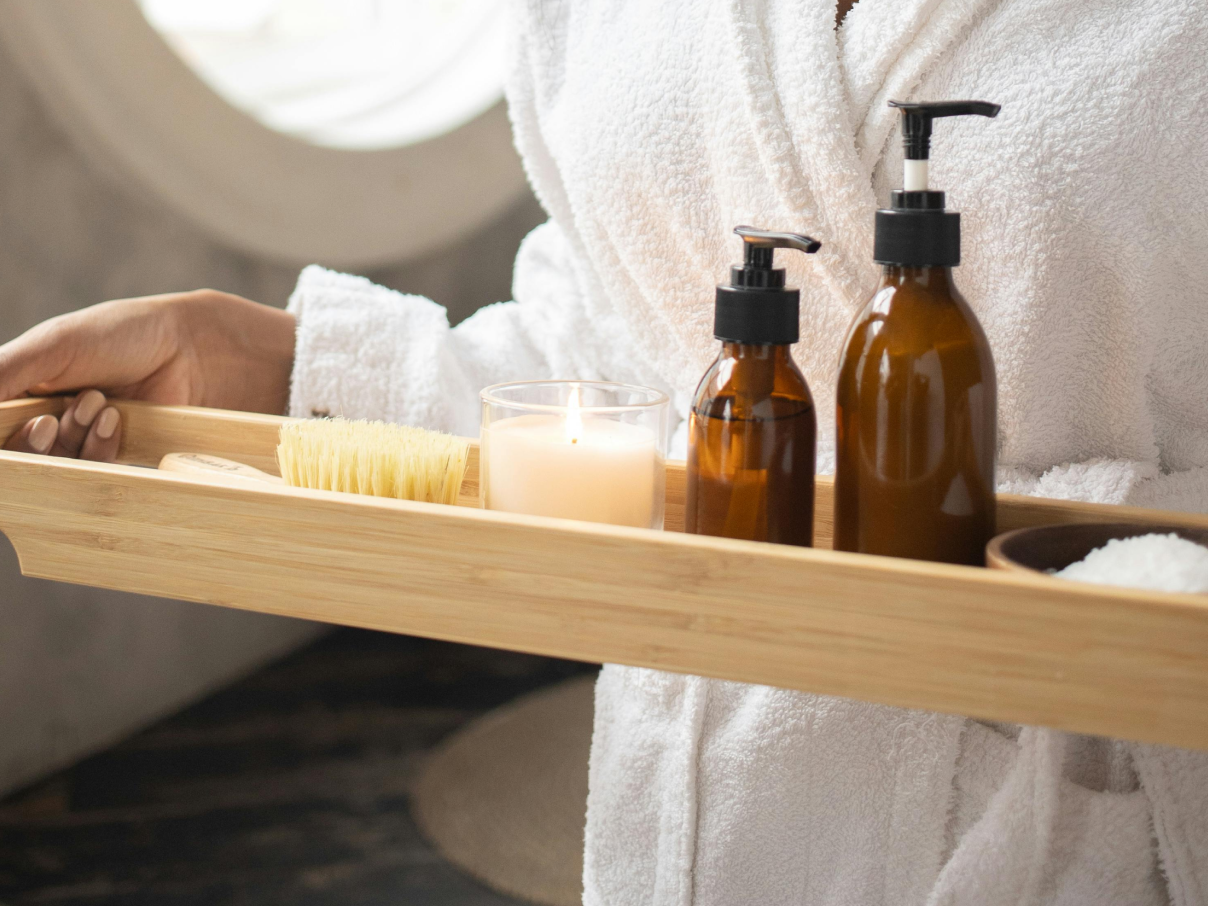 Spa amenities on a wooden tray with a candle and bottles, held by a person in a robe.