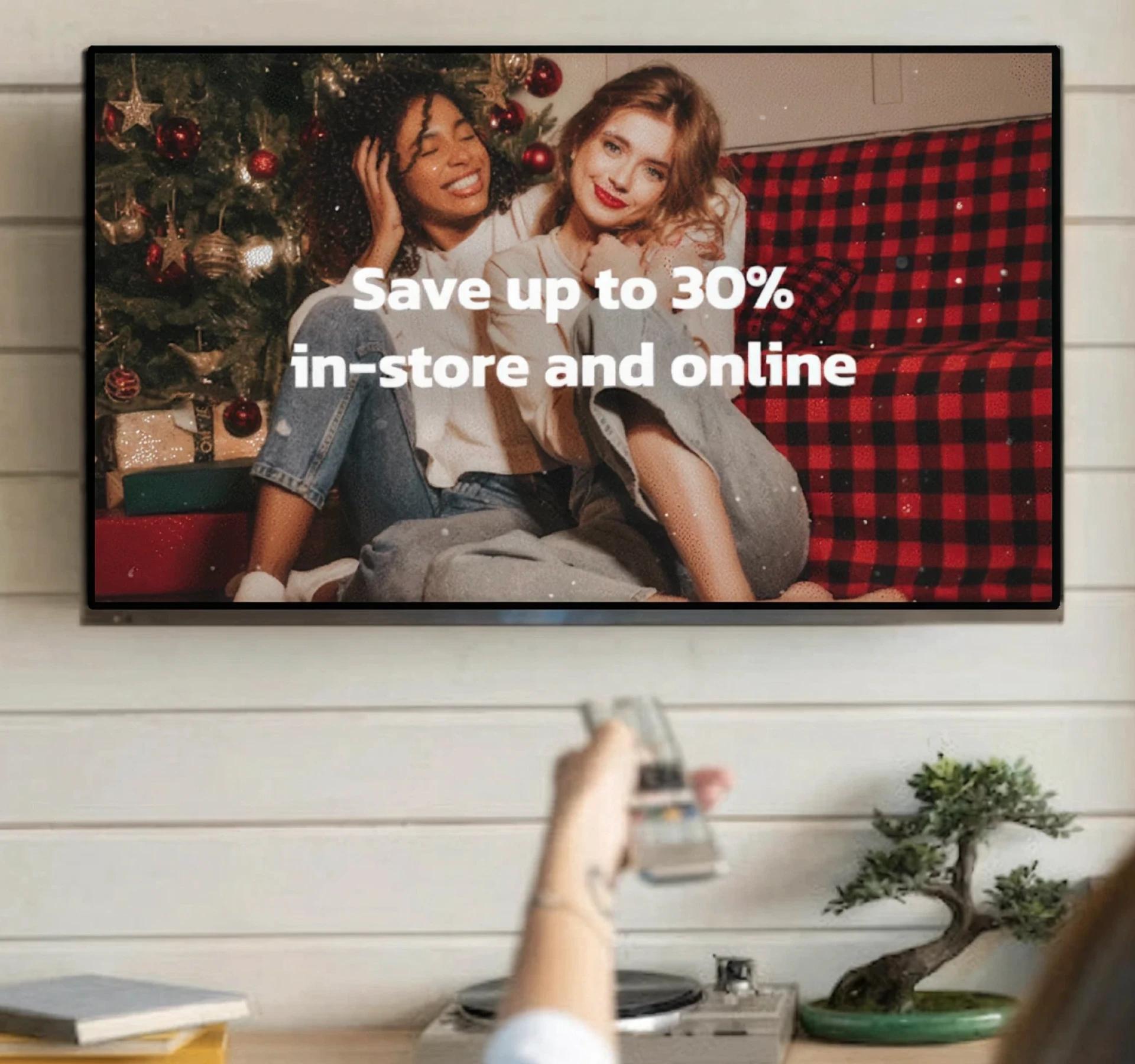 Person pointing a remote at a TV displaying a holiday sale ad with two people beside a Christmas tree.